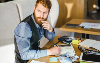 A bearded man in a blue shirt and black vest sits at a table filled with papers, photos, post-it notes, and electronic devices. He looks thoughtful, resting his hand on his chin as he holds a pen over an open notebook.