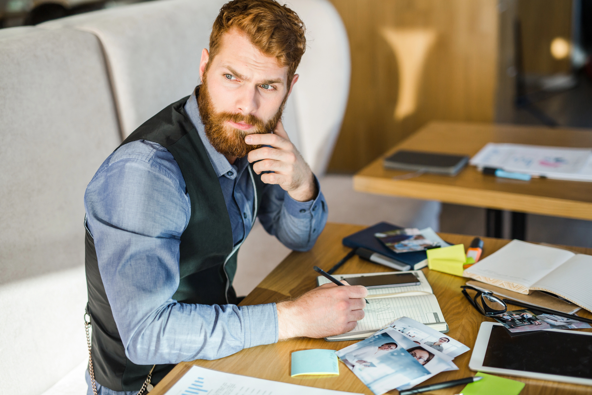 A bearded man in a blue shirt and black vest sits at a table filled with papers, photos, post-it notes, and electronic devices. He looks thoughtful, resting his hand on his chin as he holds a pen over an open notebook.