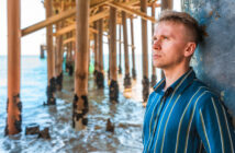 A person wearing a striped shirt leans against a pillar under a pier. They gaze thoughtfully towards the ocean. The sunlit sea and wooden pier structure are visible in the background.