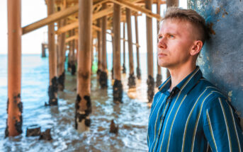 A person wearing a striped shirt leans against a pillar under a pier. They gaze thoughtfully towards the ocean. The sunlit sea and wooden pier structure are visible in the background.