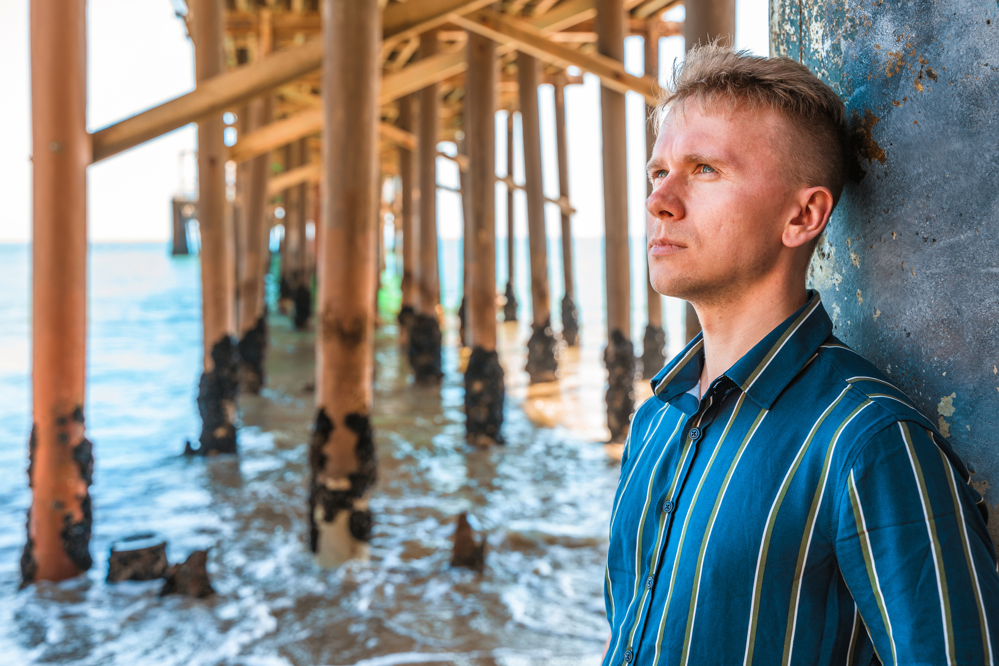 A person wearing a striped shirt leans against a pillar under a pier. They gaze thoughtfully towards the ocean. The sunlit sea and wooden pier structure are visible in the background.