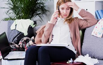 A woman sits on a cluttered sofa, talking on the phone with a worried expression. She holds paperwork on her lap and a laptop nearby. Baby clothes and a pacifier are scattered around, indicating a busy home environment.