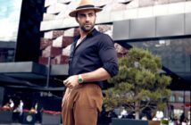 A stylish man in a black shirt and brown pants stands confidently outdoors. He wears a tan fedora and a green watch, holding sunglasses. A modern building and a small tree are in the background.