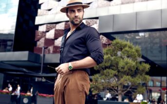 A stylish man in a black shirt and brown pants stands confidently outdoors. He wears a tan fedora and a green watch, holding sunglasses. A modern building and a small tree are in the background.