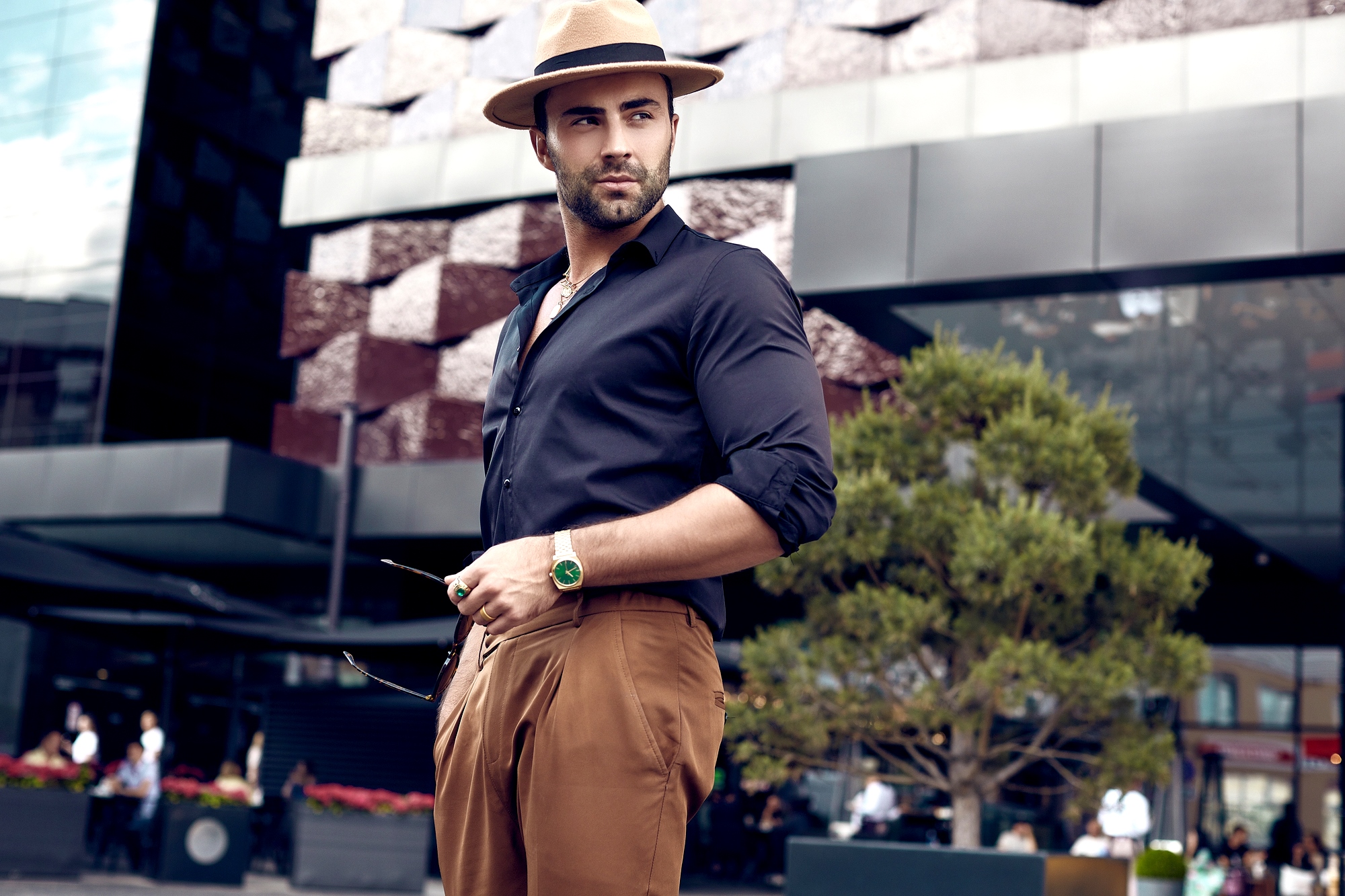 A stylish man in a black shirt and brown pants stands confidently outdoors. He wears a tan fedora and a green watch, holding sunglasses. A modern building and a small tree are in the background.