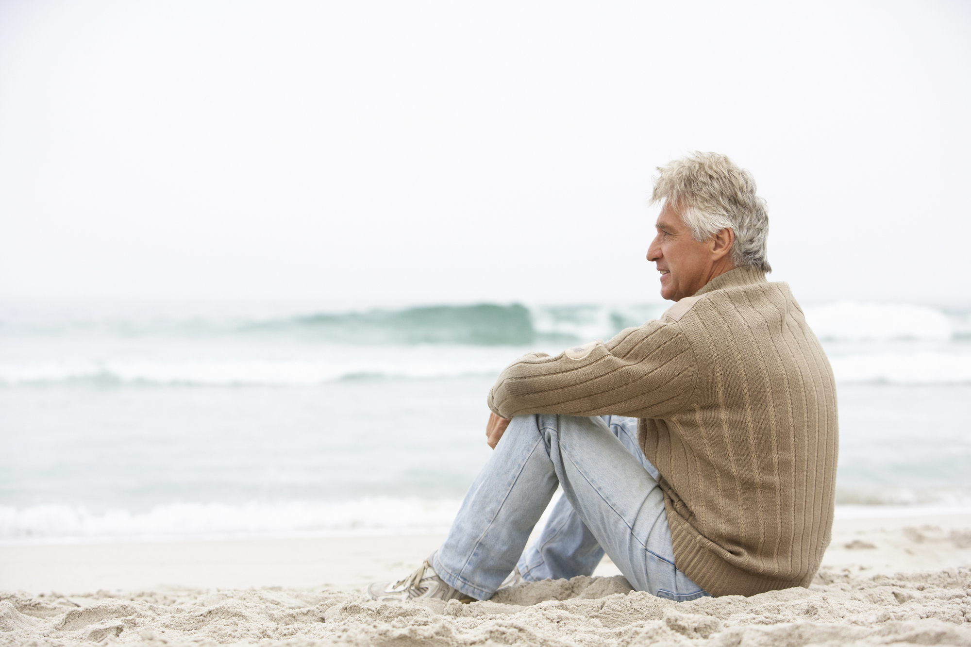 A man with gray hair sits on a sandy beach, facing the sea. He is wearing a brown knitted sweater and light blue jeans. The waves are gently rolling in the background, and the sky is overcast. The man appears to be relaxed and contemplative.