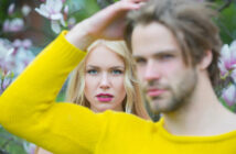 A woman with long blonde hair and pink lipstick stands in focus amidst blooming flowers, while a man with light brown hair wearing a yellow sweater stands out of focus in the foreground.