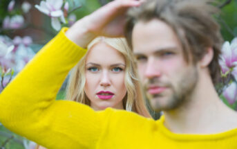 A woman with long blonde hair and pink lipstick stands in focus amidst blooming flowers, while a man with light brown hair wearing a yellow sweater stands out of focus in the foreground.