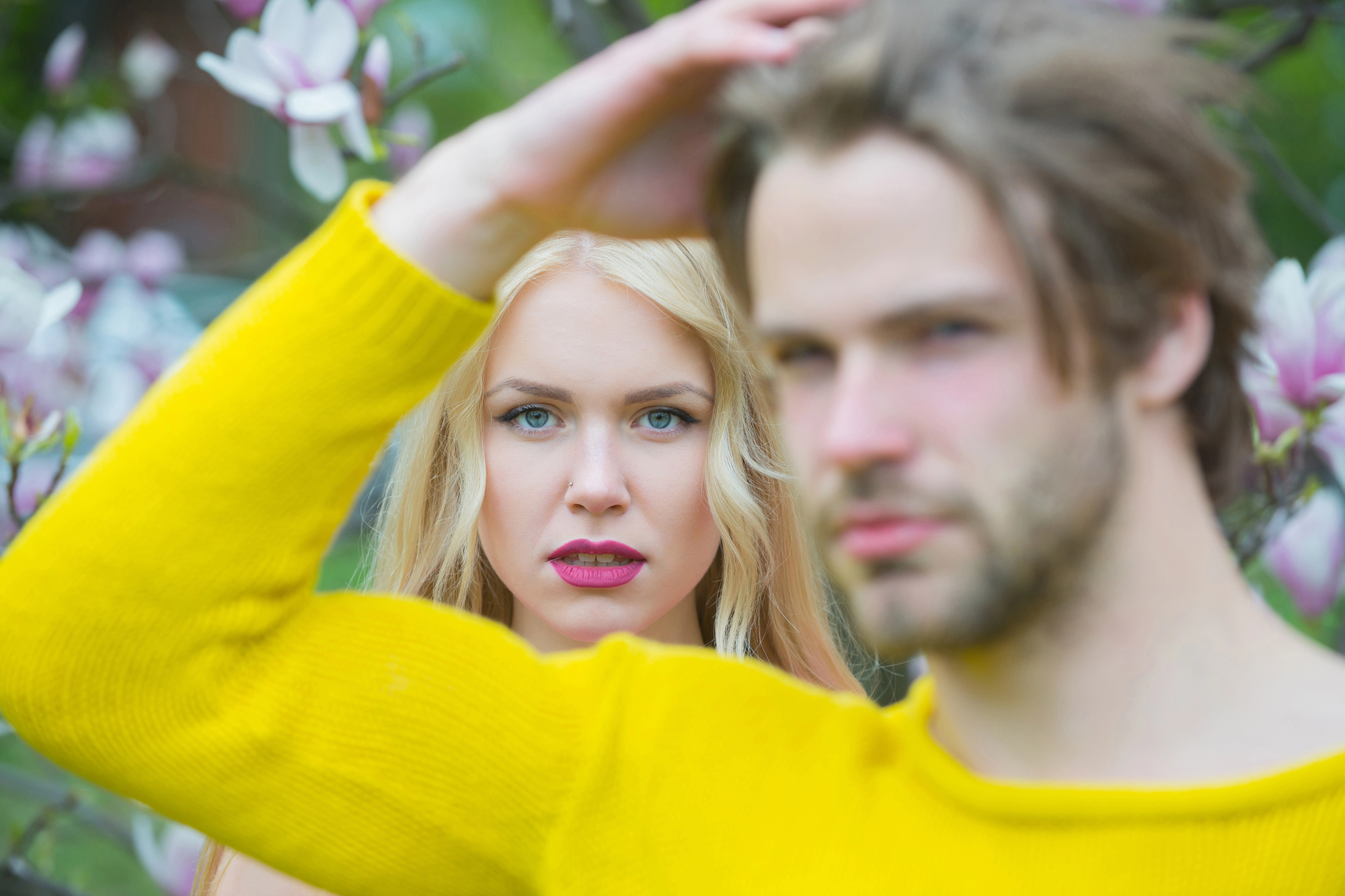 A woman with long blonde hair and pink lipstick stands in focus amidst blooming flowers, while a man with light brown hair wearing a yellow sweater stands out of focus in the foreground.