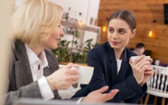 Two women are sitting at a table in a café, holding coffee cups and engaged in conversation. One woman has blonde hair and a gray suit, while the other has brown hair and a dark blazer. The background features wooden walls and soft lighting.