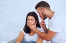 A man with a beard puts a finger to his lips, gesturing for silence, while placing his other hand over a woman's mouth. The woman looks worried and thoughtful. Both are wearing white shirts, and the background is a soft, neutral color.