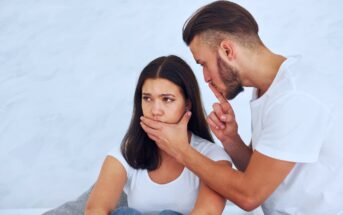 A man with a beard puts a finger to his lips, gesturing for silence, while placing his other hand over a woman's mouth. The woman looks worried and thoughtful. Both are wearing white shirts, and the background is a soft, neutral color.