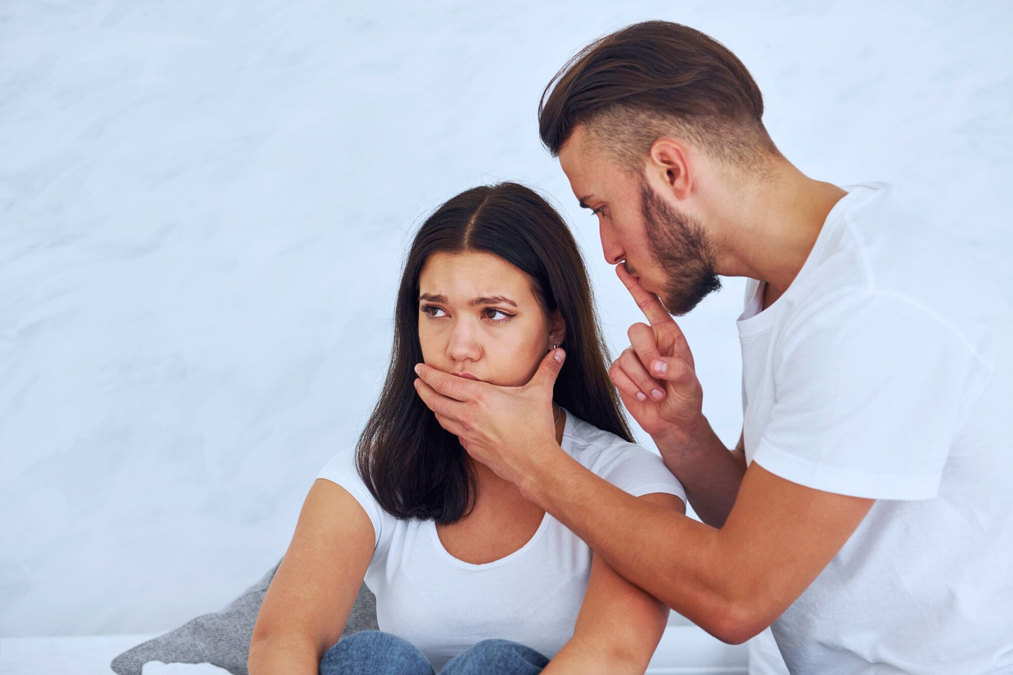 A man with a beard puts a finger to his lips, gesturing for silence, while placing his other hand over a woman's mouth. The woman looks worried and thoughtful. Both are wearing white shirts, and the background is a soft, neutral color.