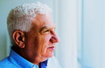 An older man with short white hair is looking thoughtfully out of a window. He is wearing a blue collared shirt. The background is softly blurred, suggesting daylight coming through the window.