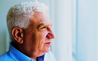 An older man with short white hair is looking thoughtfully out of a window. He is wearing a blue collared shirt. The background is softly blurred, suggesting daylight coming through the window.