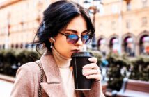 A woman wearing sunglasses and a brown coat sips from a black takeaway coffee cup. She has dark hair and is standing outdoors with a blurred background featuring buildings and greenery.