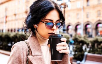 A woman wearing sunglasses and a brown coat sips from a black takeaway coffee cup. She has dark hair and is standing outdoors with a blurred background featuring buildings and greenery.