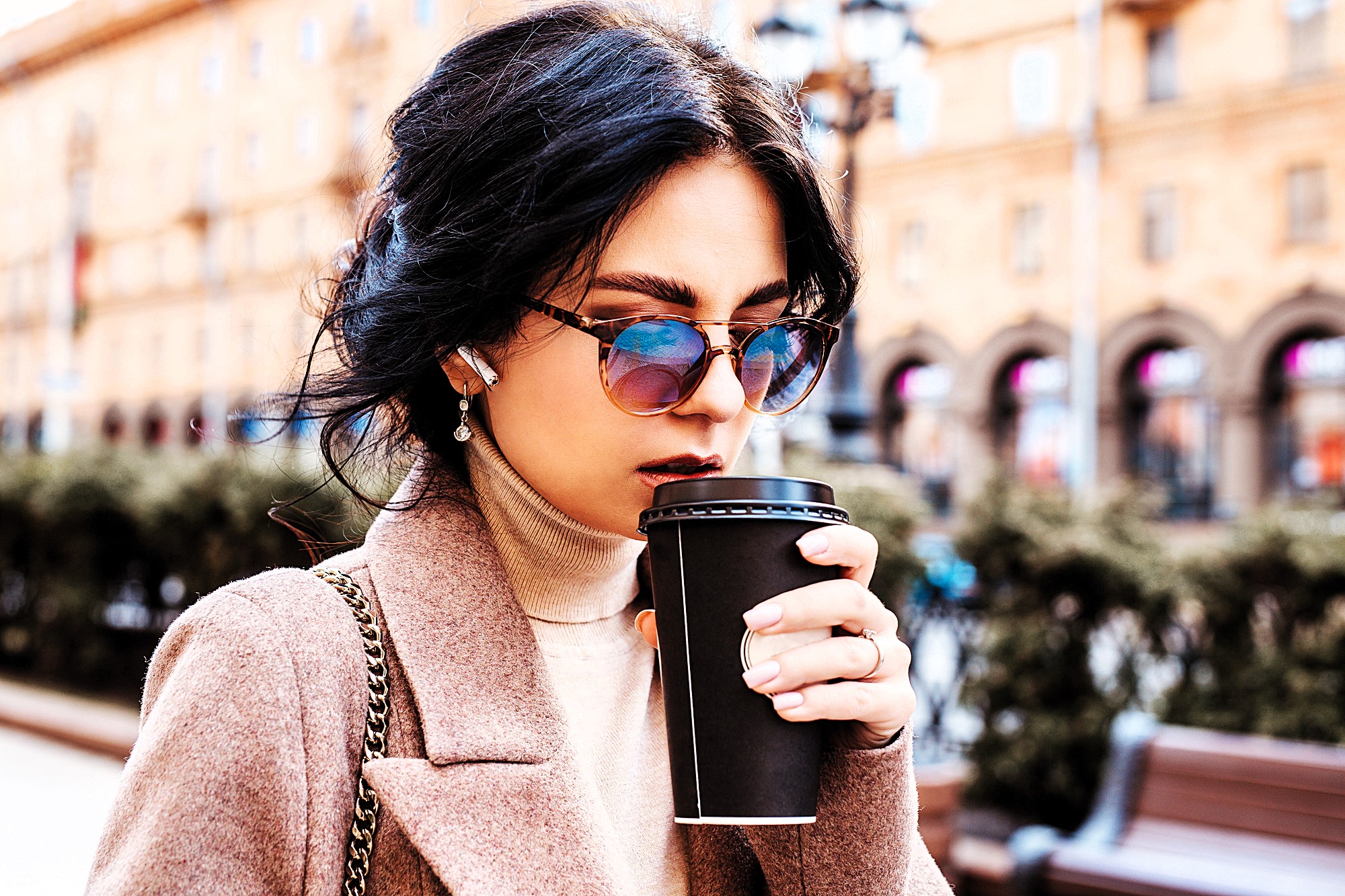 A woman wearing sunglasses and a brown coat sips from a black takeaway coffee cup. She has dark hair and is standing outdoors with a blurred background featuring buildings and greenery.