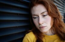 A woman with long red hair and freckles leans against a dark, ridged wall, wearing a yellow top. She appears pensive, gazing downward with a neutral expression.