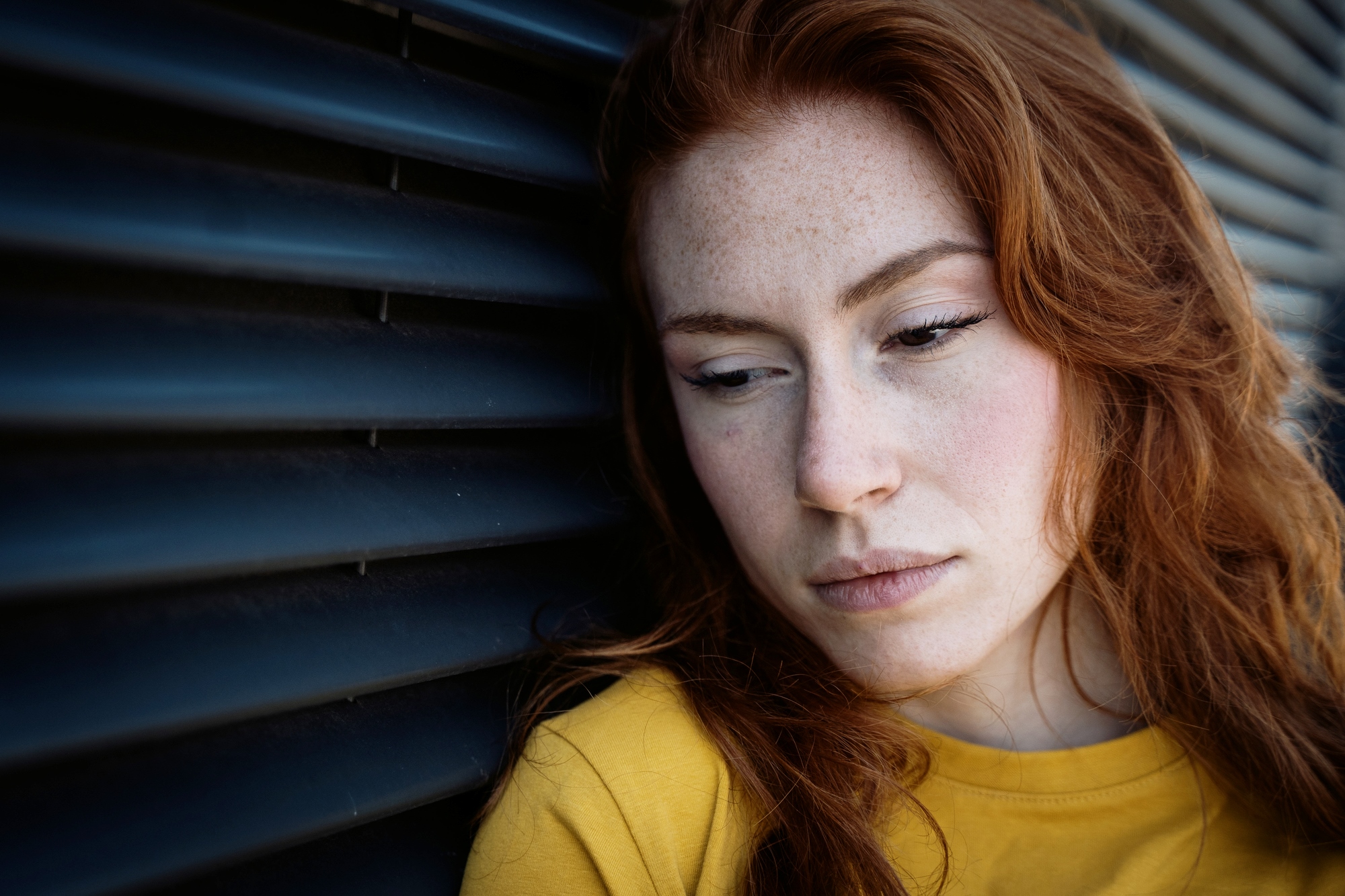 A woman with long red hair and freckles leans against a dark, ridged wall, wearing a yellow top. She appears pensive, gazing downward with a neutral expression.