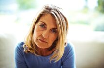 A woman with long blonde hair sits indoors, wearing a blue top. She has a contemplative expression, with natural light softly illuminating her face. The background is blurred, suggesting a serene and calm environment.