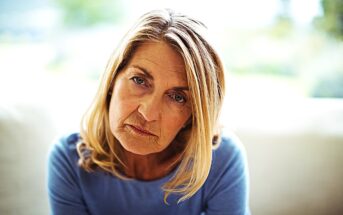 A woman with long blonde hair sits indoors, wearing a blue top. She has a contemplative expression, with natural light softly illuminating her face. The background is blurred, suggesting a serene and calm environment.