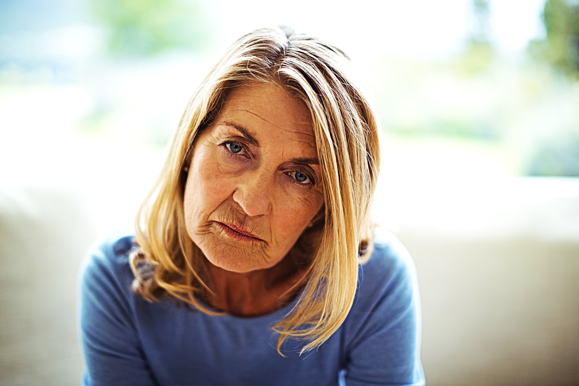 A woman with long blonde hair sits indoors, wearing a blue top. She has a contemplative expression, with natural light softly illuminating her face. The background is blurred, suggesting a serene and calm environment.