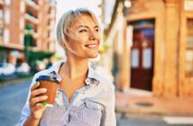 A person with short blonde hair holds a coffee cup and smiles while walking on a city street. They're wearing a blue striped shirt, and the background features buildings and blurred greenery, suggesting a sunny day.