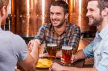 Three men sitting at a brewery table, holding glasses of beer. One man in a plaid shirt is shaking hands with another man whose back is to the camera. A bowl of chips is on the table. They are smiling with copper brewing tanks in the background.