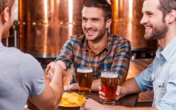 Three men sitting at a brewery table, holding glasses of beer. One man in a plaid shirt is shaking hands with another man whose back is to the camera. A bowl of chips is on the table. They are smiling with copper brewing tanks in the background.