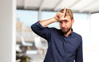 A man with a beard stands indoors, making an "L" shape with his fingers on his forehead. He is wearing a blue polka-dot shirt. The background is blurred, showing a modern office setting.