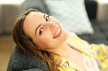 A woman with light brown hair smiles warmly while reclining on a gray sofa. She wears a yellow-striped top, and a light blue pillow is visible in the background. The setting appears bright and cozy.