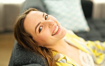 A woman with light brown hair smiles warmly while reclining on a gray sofa. She wears a yellow-striped top, and a light blue pillow is visible in the background. The setting appears bright and cozy.