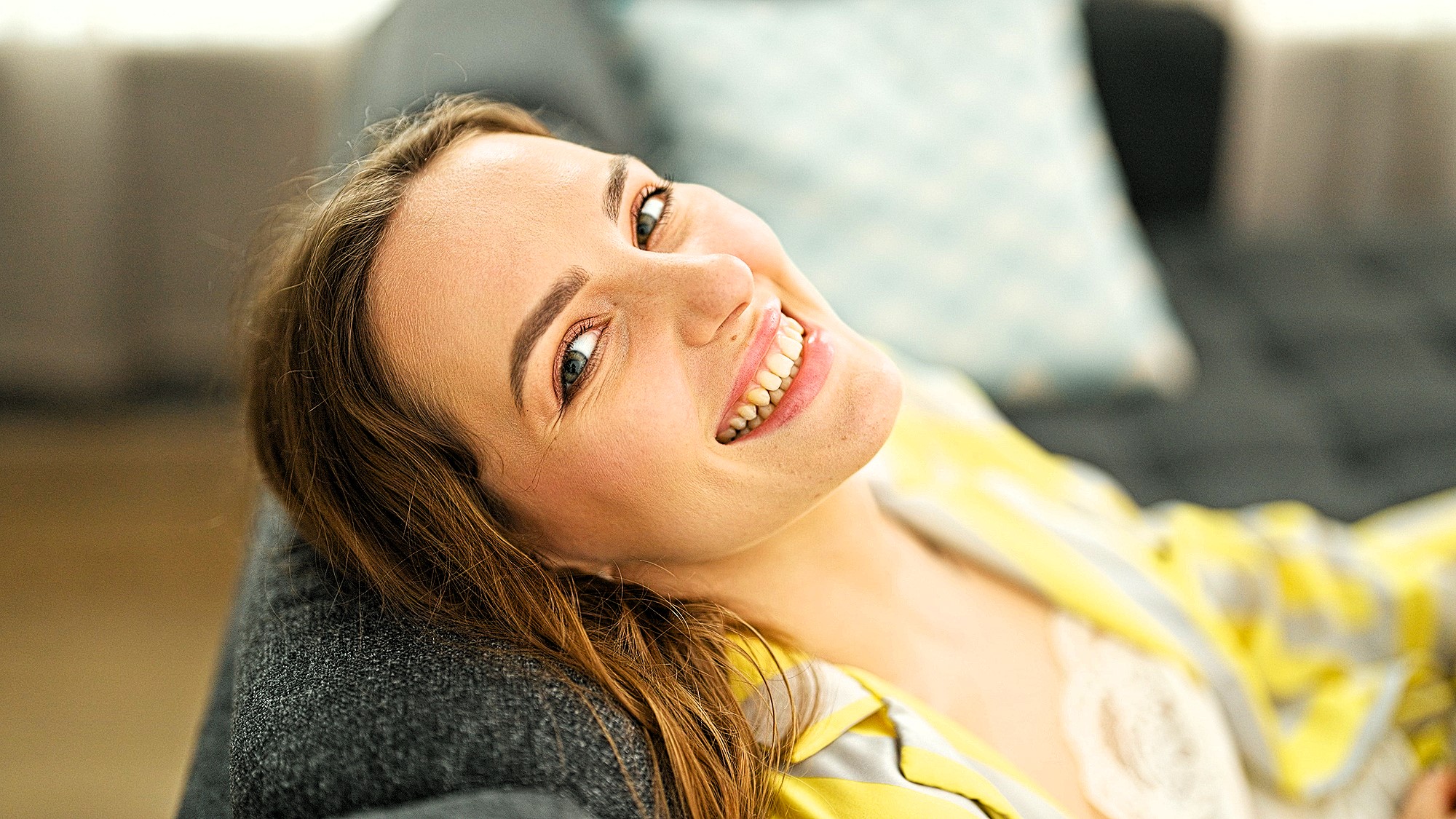 A woman with light brown hair smiles warmly while reclining on a gray sofa. She wears a yellow-striped top, and a light blue pillow is visible in the background. The setting appears bright and cozy.