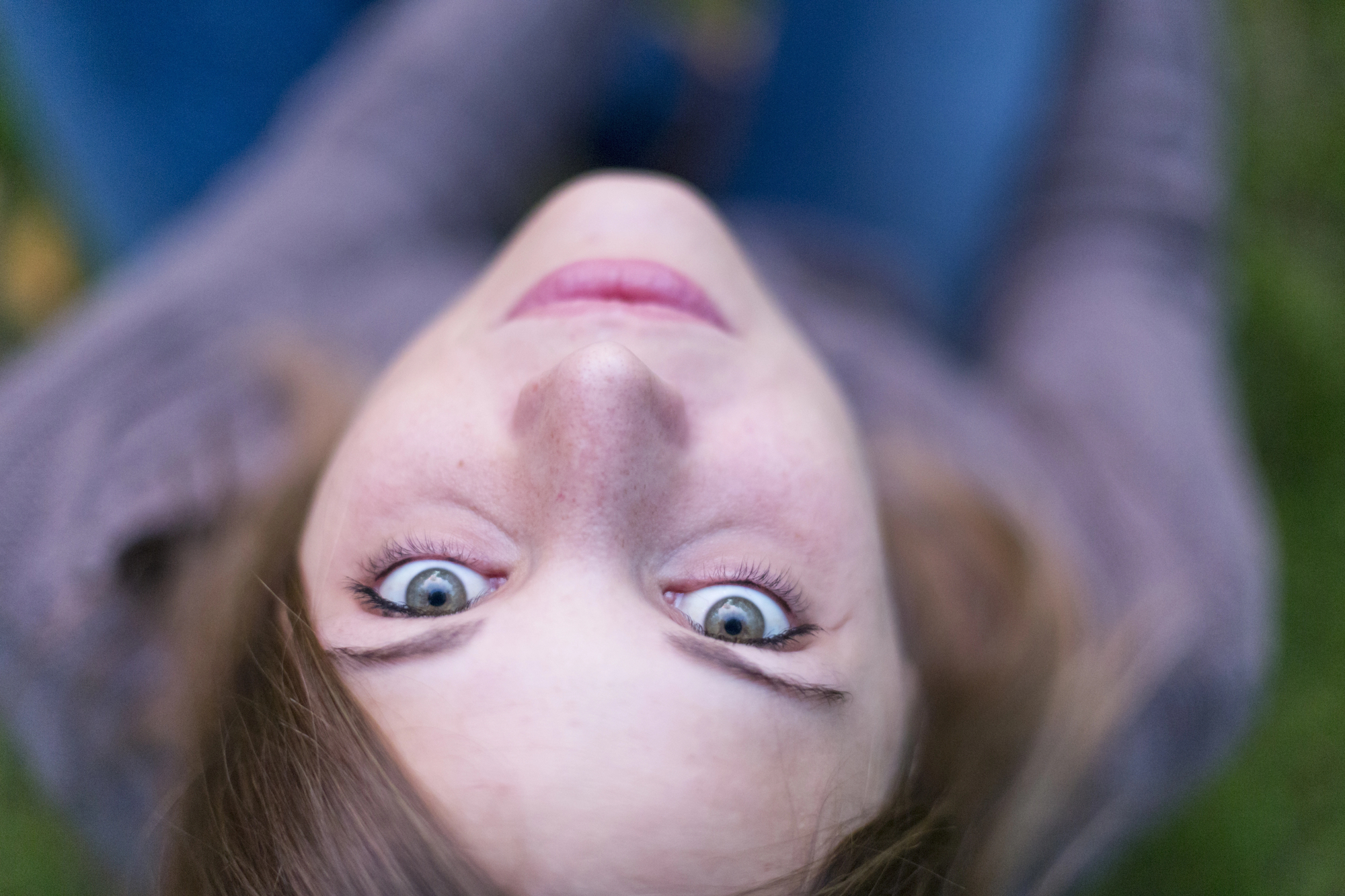 Close-up portrait of a person with blue eyes, looking directly at the camera. The photo is taken from an overhead angle, capturing their face and part of their brown hair. They are wearing a gray top, with a blurred green background.