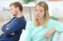 A woman wearing a light turquoise sweater looks upset, sitting on a couch with her arm draped over the back. A man in a blue sweater sits beside her, turned away with his arms crossed. The background is softly focused.