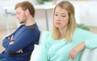 A woman wearing a light turquoise sweater looks upset, sitting on a couch with her arm draped over the back. A man in a blue sweater sits beside her, turned away with his arms crossed. The background is softly focused.