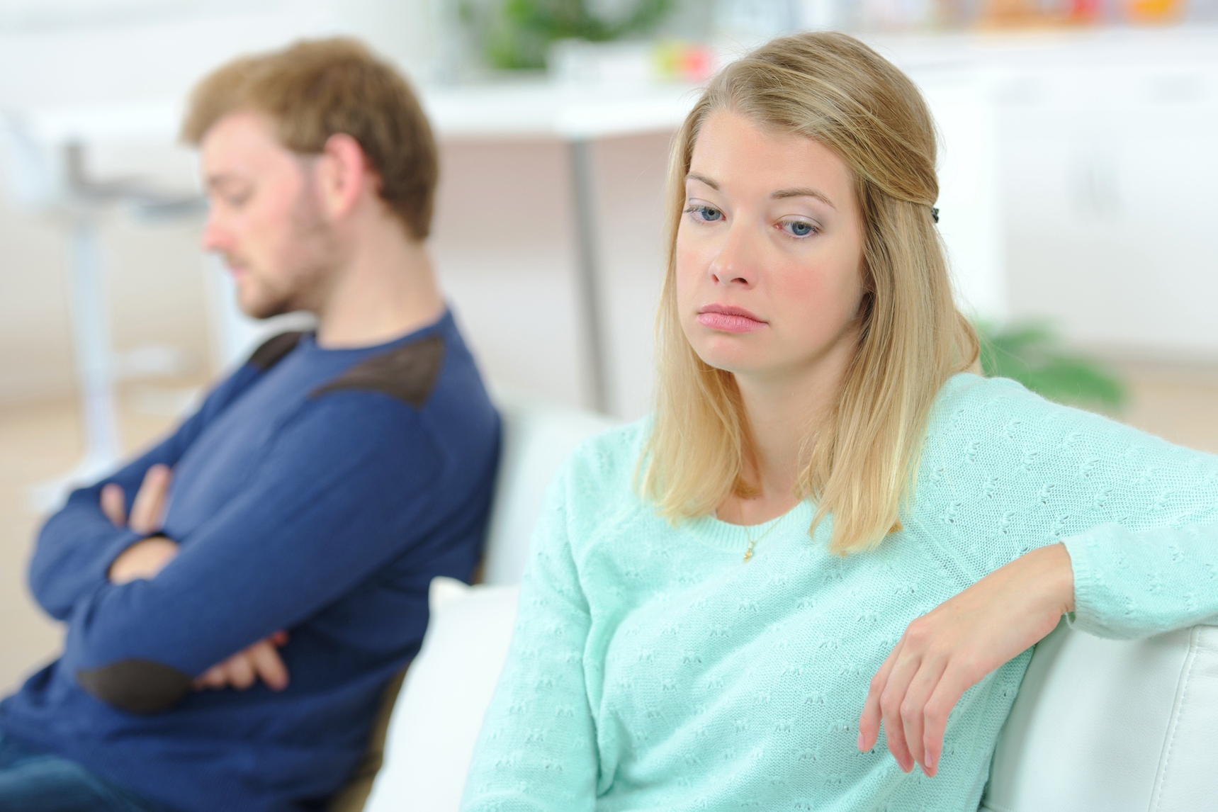 A woman wearing a light turquoise sweater looks upset, sitting on a couch with her arm draped over the back. A man in a blue sweater sits beside her, turned away with his arms crossed. The background is softly focused.