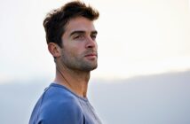 A man with short dark hair and a slight beard gazes into the distance. He is wearing a light blue sweater, standing against a soft, blurred background that hints at a serene, overcast sky.