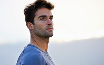 A man with short dark hair and a slight beard gazes into the distance. He is wearing a light blue sweater, standing against a soft, blurred background that hints at a serene, overcast sky.