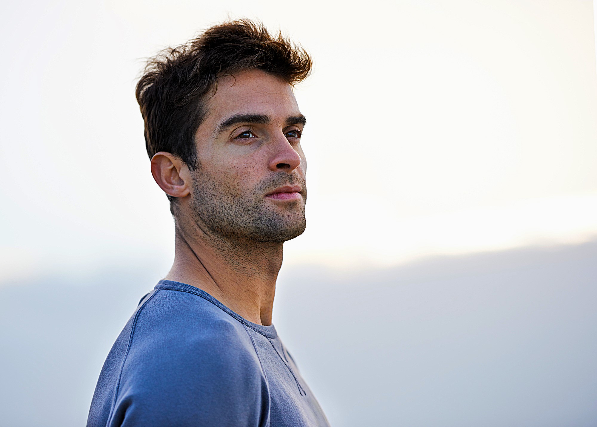 A man with short dark hair and a slight beard gazes into the distance. He is wearing a light blue sweater, standing against a soft, blurred background that hints at a serene, overcast sky.