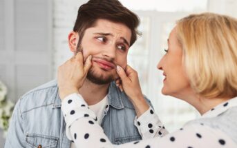 A woman playfully pinches the cheeks of a young man with a beard. Both are indoors and smiling. The woman is wearing a polka dot blouse, and the man has a denim jacket. The scene suggests a lighthearted interaction.