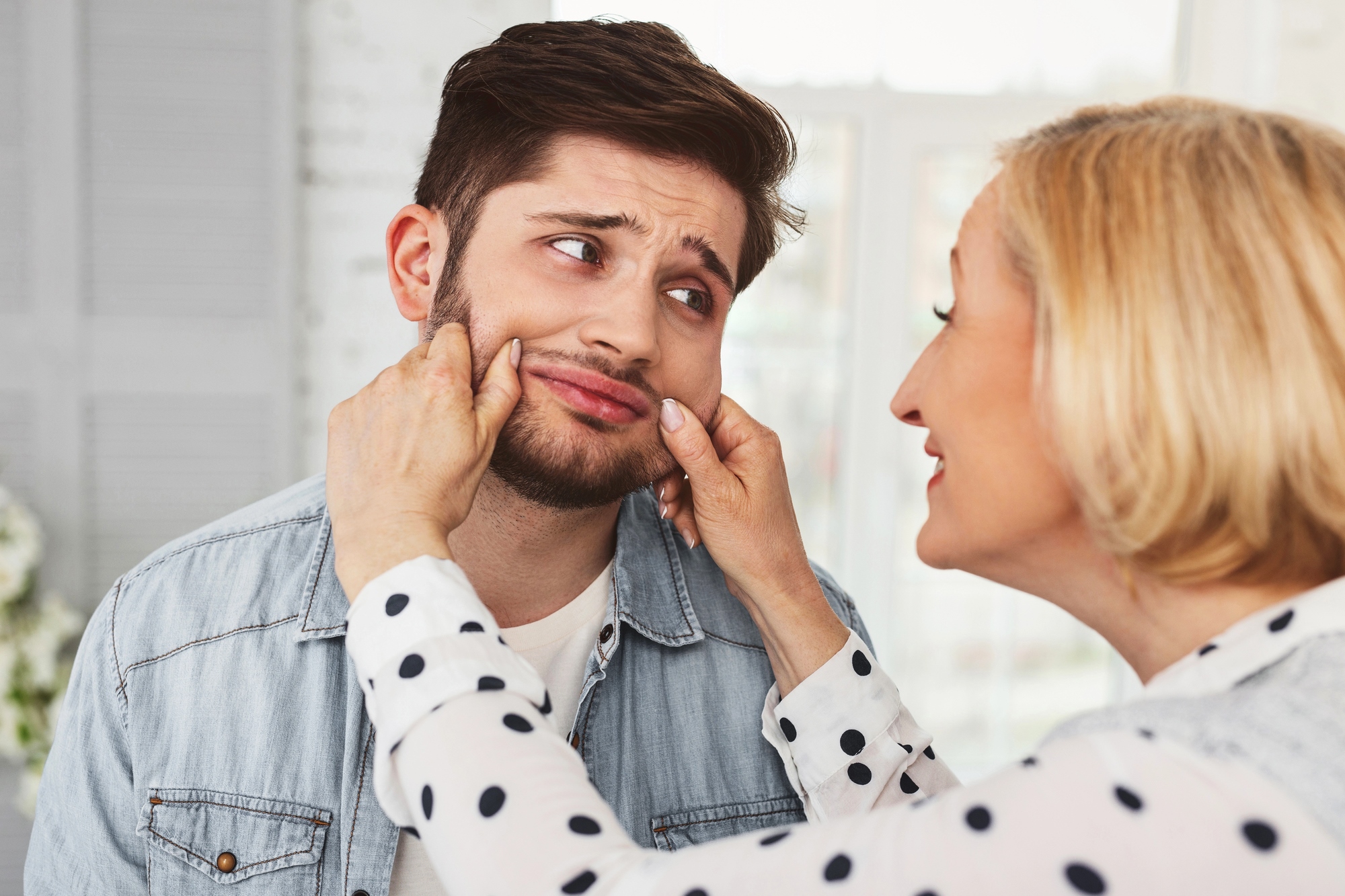 A woman playfully pinches the cheeks of a young man with a beard. Both are indoors and smiling. The woman is wearing a polka dot blouse, and the man has a denim jacket. The scene suggests a lighthearted interaction.