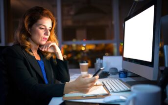 A woman in an office is illuminated by a computer screen while holding a smartphone. She is seated at a desk with a keyboard, coffee cup, and stationery. The background shows city lights through a window, indicating nighttime.