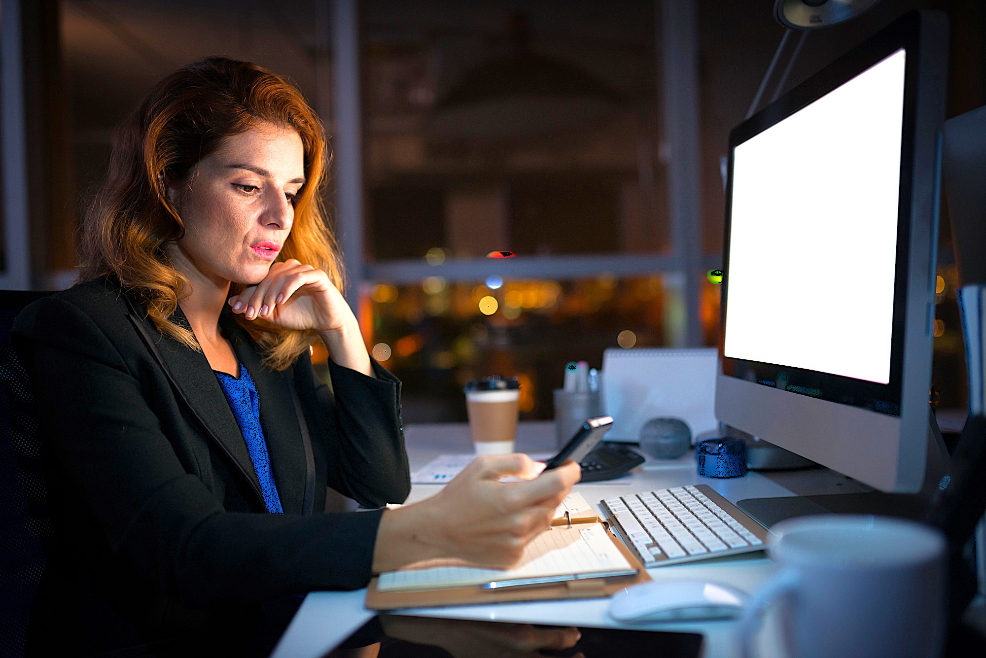 A woman in an office is illuminated by a computer screen while holding a smartphone. She is seated at a desk with a keyboard, coffee cup, and stationery. The background shows city lights through a window, indicating nighttime.