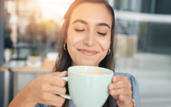 A woman with long hair is smiling with her eyes closed, holding a large mug of coffee with both hands. She seems to be savoring the aroma. The background is softly blurred, suggesting a cozy, relaxed atmosphere.