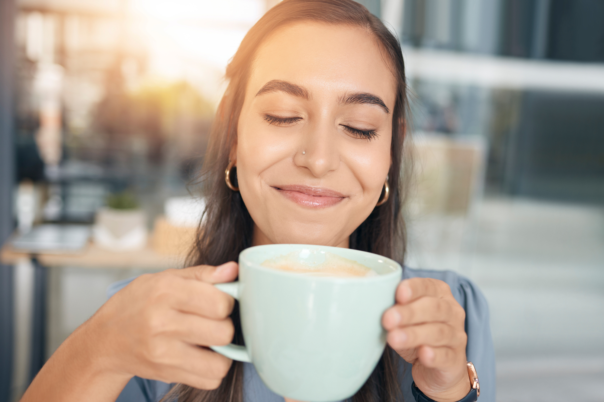 A woman with long hair is smiling with her eyes closed, holding a large mug of coffee with both hands. She seems to be savoring the aroma. The background is softly blurred, suggesting a cozy, relaxed atmosphere.