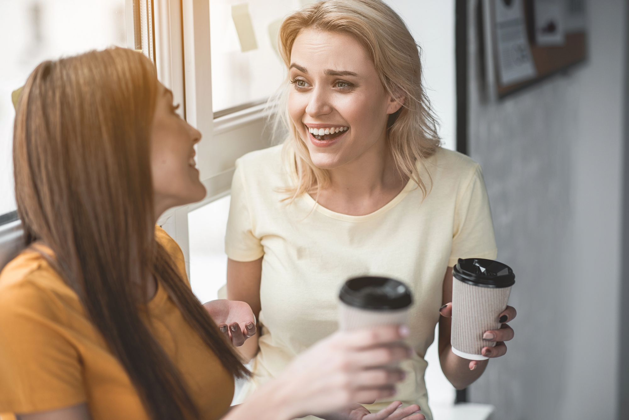 Two women are sitting by a window, smiling and talking while holding disposable coffee cups. They appear to be enjoying a casual conversation, dressed in light-colored tops, with natural light streaming in from outside.