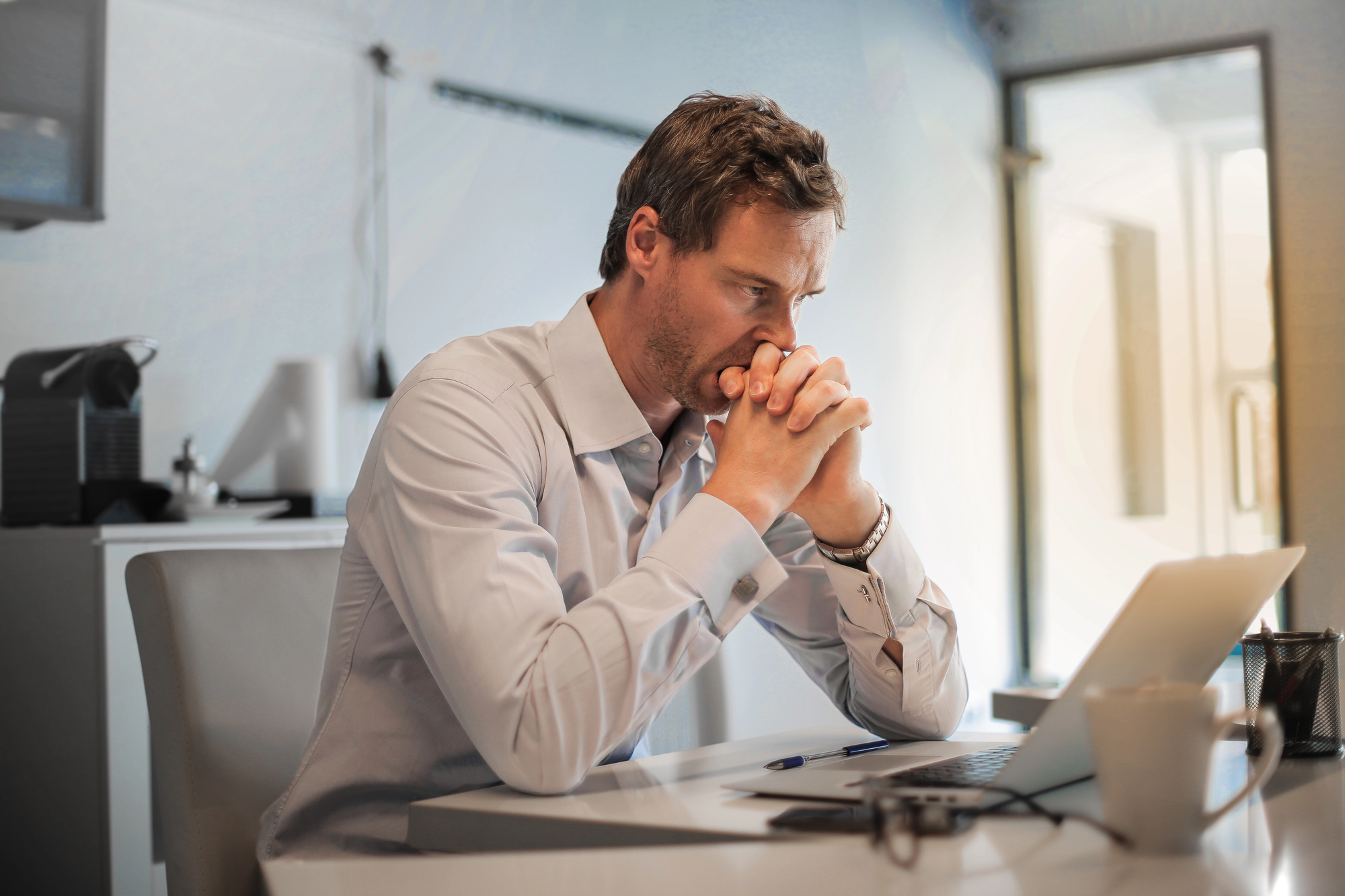 A man in a white shirt sits at a desk, focused intently on a laptop screen. His hands are clasped in front of his face, conveying deep thought. The desk has a mug, glasses, a pen, and notepad. The room is well-lit with natural light.