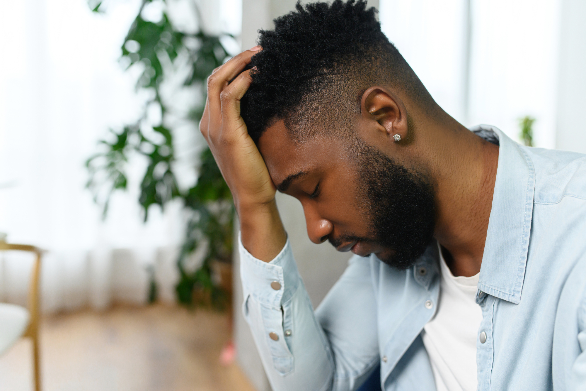 A man with a beard and short curly hair wearing a light blue shirt and a white T-shirt sits with his head in his hand, appearing stressed or deep in thought. The background includes blurred elements of indoor plants and furniture.